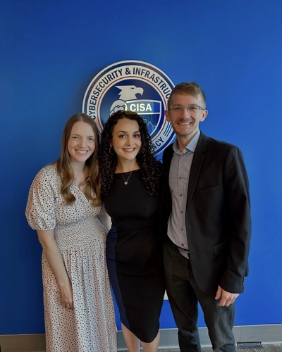 Rina Rakipi, flanked by Karissa Chabot-Purchase and Max Olivero pose together for a photo in front of a solid blue wall with a circular CISA agency logo seal.