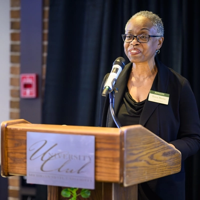 A woman with short grey hair and glasses, identified by her name tag as Teresa Mastin, speaking into a microphone at a wooden podium. She is wearing a black blazer and top. The podium features a silver "University Club" plaque with "Michigan State University" visible. She is standing in front of a dark curtain backdrop with a brick pillar to the left.