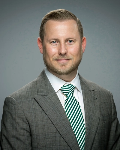 A man in a charcoal blazer with a light plaid, over a white button-down and a green and white striped tie in front of a gray background.