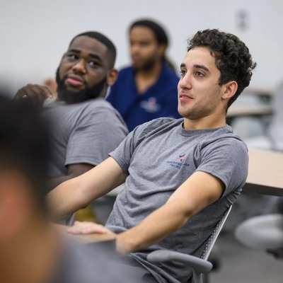 a young adult male in a grey tshirt sits at a desk during a seminar. Another young man also in a grey tshirt looks on.