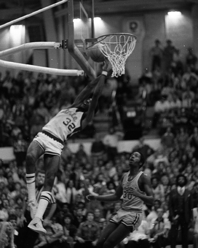 Black-and-white photo of a basketball game showing a player wearing jersey number 32 jumping high to dunk the ball into the hoop, with another player nearby watching and a large crowd seated in the background of an indoor gymnasium.