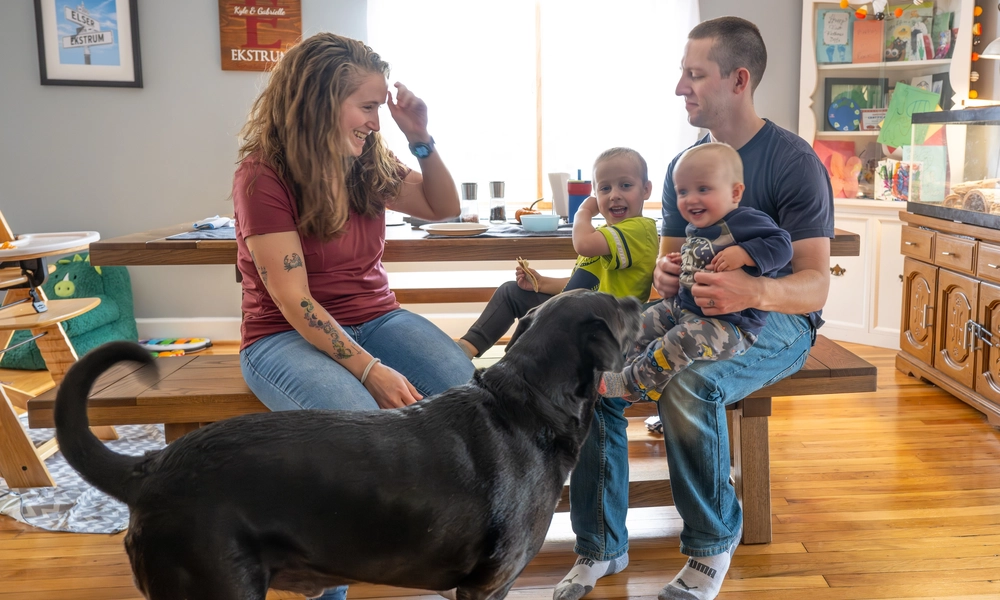 Ekstrum family sits together in dining room with their dog as parents and children smile and play.