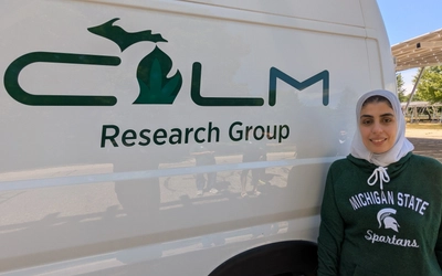 Omayma Alshaarawy, associate professor of family medicine at Michigan State University, stands beside a vehicle displaying the CALM Research Group logo. She is wearing a green Michigan State Spartans sweatshirt and a white headscarf, smiling outdoors on a sunny day with solar panel structures visible in the background.
