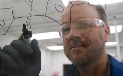 Researcher wears protective eyewear writing a chemical structure on glass inside a laboratory.