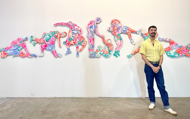 Artist Gustavo Uriel Ayala stands in front of his panoramic artwork entry for ArtPrize depicting sequential, vibrantly colored figures engaged in dynamic wrestling holds, maneuvers, and sportsmanship against a plain background.