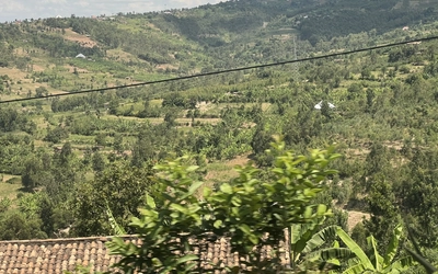 A scenic view of rolling green hills covered with farms, trees and scattered homes under a partly cloudy sky. The rural agricultural landscape features cultivated fields stretching across the hillsides.