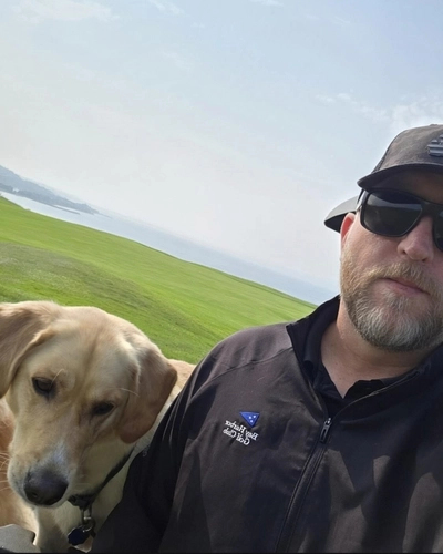 Selfie of Christopher Burnett, wearing a black hat and sunglasses with a yellow lab and Lake MIchigan in the background at Bay Harbor Golf Club