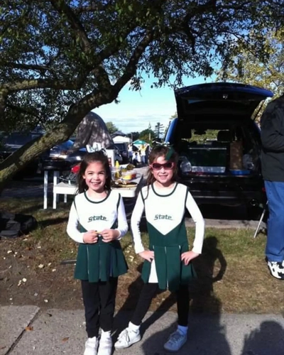 Two young girls in green and white cheer uniforms smile at an MSU tailgate, standing in front of the family’s tailgate setup on game day.