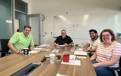 Four individuals smile while sitting around a wooden conference table with the board game Connected Community laid out on the table.