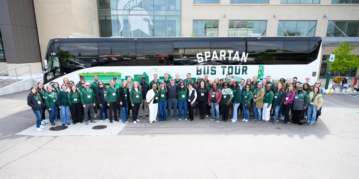 Large group of people pose in front of a bus with SPARTAN BUS TOUR label on it in front of a building with a Spartan helmet