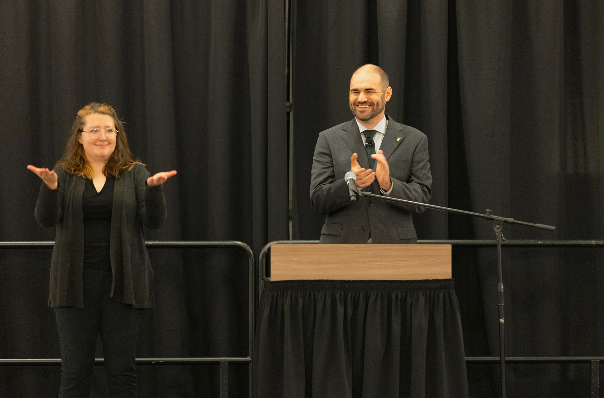 A man stands at a lectern clapping hands on stage, with an interpreter standing nearby