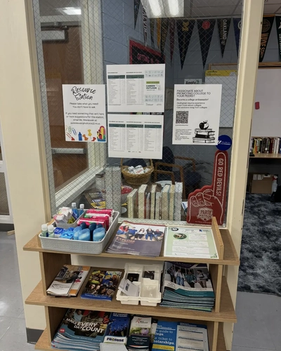A wooden three-shelf "Resource Station" located against a glass partition in a school office. The top shelf contains a grey basket filled with personal care items like deodorant, pads, and hand sanitizer. The middle and bottom shelves hold various educational brochures, college guides, and flyers. On the glass above, there are informational posters and a view into an office decorated with college pennants.