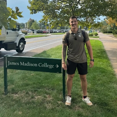 Student Colt Gray stands beside a green directional sign for James Madison College on the Michigan State University campus in East Lansing, Michigan.