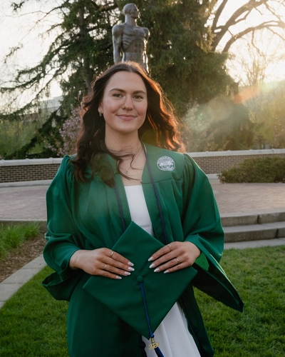 Torey Auzins wears a green graduation stole and holds a mortarboard while standing in front of the Spartan statue.