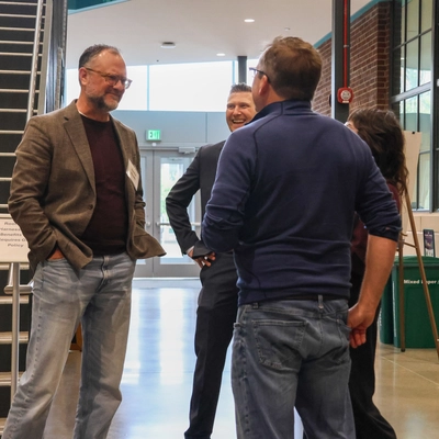 A group of four men and one woman chat casually in a bright, modern building atrium next to a large staircase and a brick wall with large windows. The man on the left wears a brown tweed blazer and jeans, smiling as he talks with a man facing away from the camera in a blue pullover. Other attendees smile in the background.
