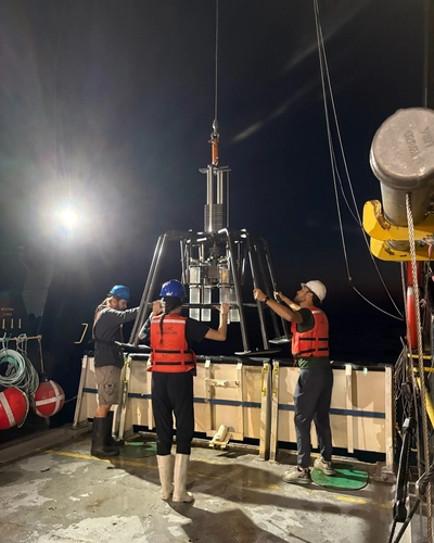 Researchers in safety vests maneuver scientific sampling equipment on a ship deck at night.