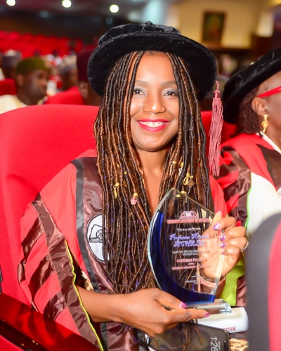 Nwando Achebe in red and burgundy robe holds up a glass award in an auditorium setting.