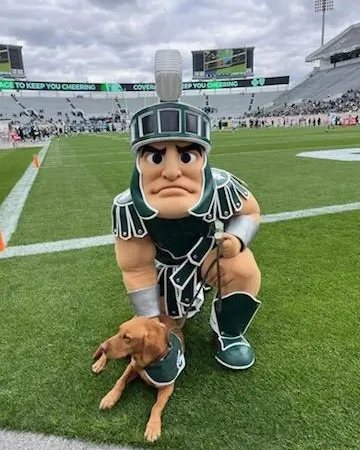 Sparty mascot kneeling next to Zeke the Wonder Dog on the football field at Spartan Stadium.