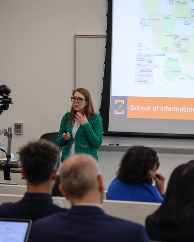 A woman with long auburn hair and glasses wearing a green textured blazer speaks at the front of a lecture hall. Behind her, a projector screen displays a map of the United States and text reading "School of Information". The backs of audience members' heads are visible in the foreground