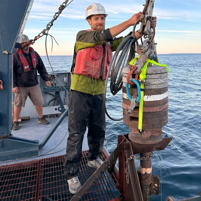 Dalton Hardisty stands on a metal grate deck of a boat on water with marine research equipment wearing a hard hat and bright green jacket beneath a red life vest.