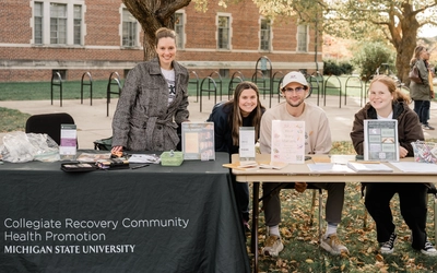 Four people staffing a Michigan State University Collegiate Recovery Community table at an outdoor fall festival.