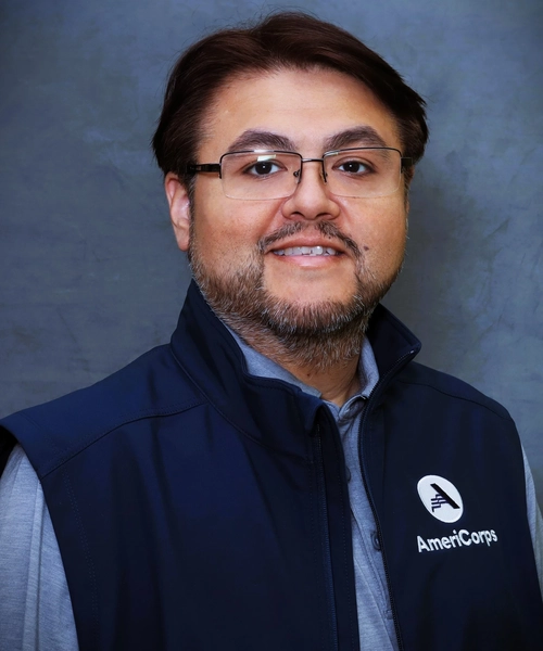 A headshot of a man with dark hair, glasses, and a groomed beard smiling at the camera. He is wearing a grey collared shirt under a dark blue vest that has the AmeriCorps logo on the left side. The background is a solid, textured grey.