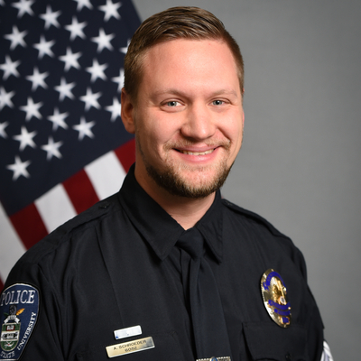 Aaron Schroeder, a uniformed Michigan State University police officer smiling in front of an American flag.