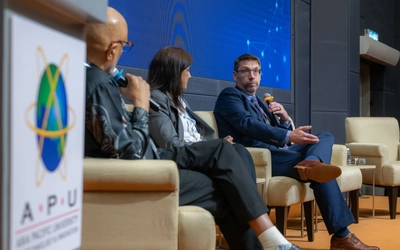 Three people sit on a conference stage as part of a panel presentation. A podium is in the foreground and there is a blue background behind them.