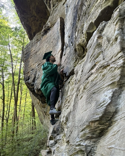 A man rock climbs wearing a graduation cap and gown