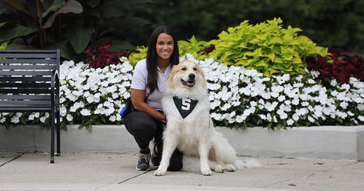 A woman kneels beside a large white and tan fluffy dog wearing a green bandana with a white "S" on it. They are sitting on a sidewalk in front of a flower bed filled with white, red, and green plants. Both are smiling, and the scene appears bright and cheerful outdoors.