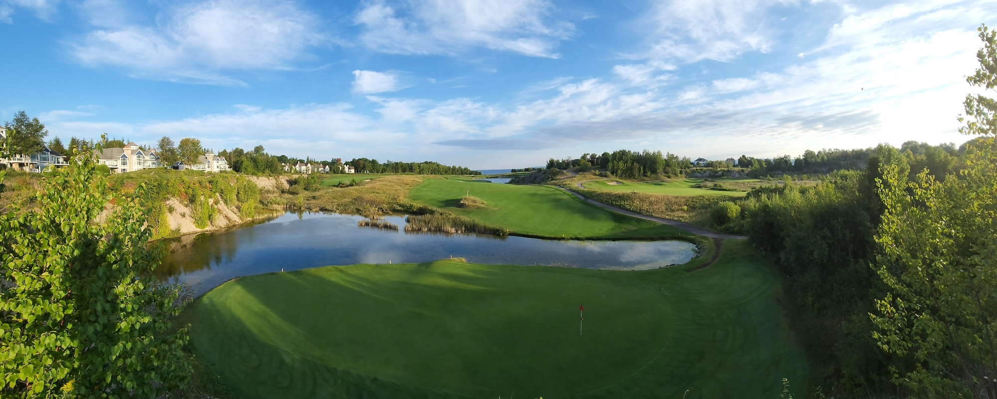 Panoramic view of a golf hole at Bay Harbor Golf Course, with a large green, along with houses along the fairway and, in the distance, Lake Michigan