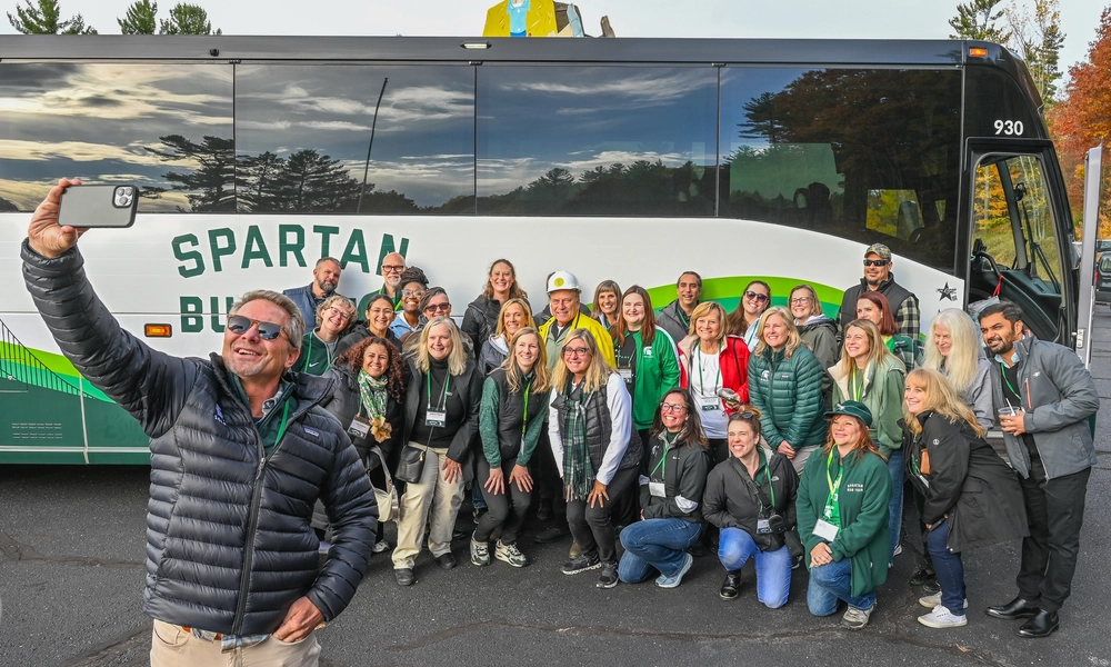 A large group of people pose for a photo in front of a white and green tour bus labeled “Spartan Bus Tour.” One person in the foreground holds up a smartphone to take a selfie with the group. The bus is parked on an asphalt lot surrounded by trees with autumn foliage, and a colorful wood figure is visible behind of the bus.