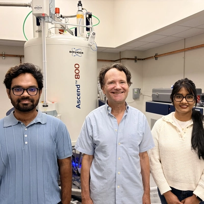 Three people stand in a laboratory in front of a large white Bruker Ascend 800 nuclear magnetic resonance (NMR) spectrometer. The individuals are positioned side by side, with scientific equipment, computers, and laboratory fixtures visible in the background.