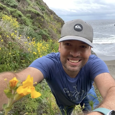 Selfie of a man climbing with yellow flowers surrounding him near the ocean.