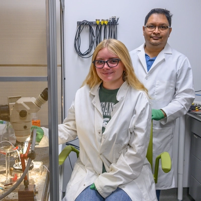 A woman sits in front beside a microscope. Behind her, a man is standing.