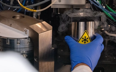 A blue-gloved hand places a metal cylinder into the interior of a cryo-electron microscope. The interior is a collection of metallic surfaces and wires. The cylinder sports a yellow and black cryogenic temperature warning