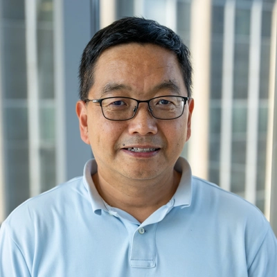 Xuefei Huang poses for a headshot in front of a window. He wears a light blue polo with an orange logo.