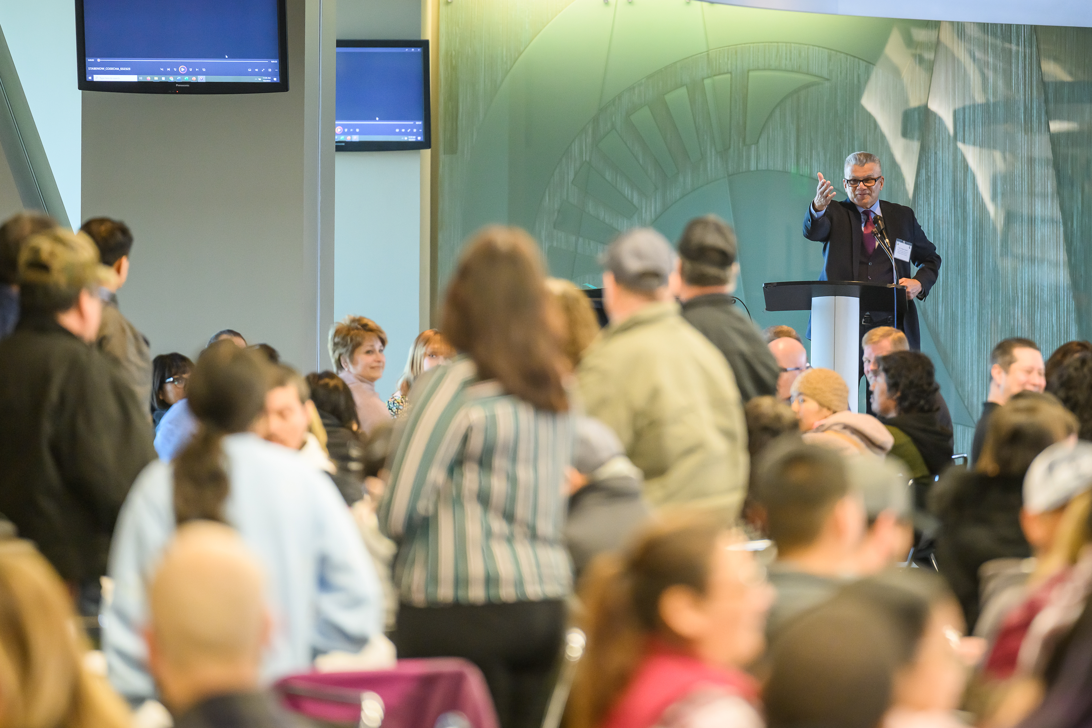 A person gestures to the audience from a lectern people are standing in the foreground