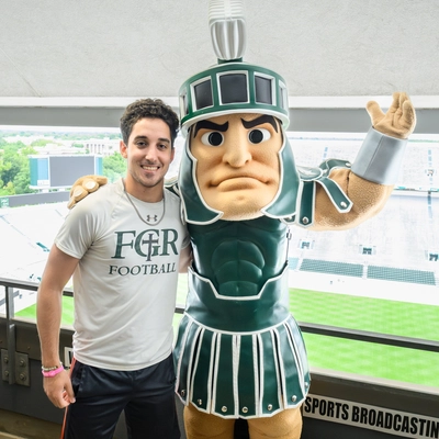 A photo of a young man wearing a cream shirt and black shorts next to Sparty the mascot