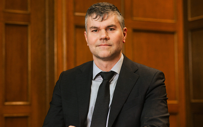 Studio portrait of a person seated in a dark suit, light dress shirt, and dark tie, facing the camera against a warm wood-paneled background. Soft lighting highlights the subject's face and upper body in a formal professional setting.