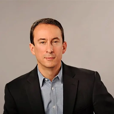 A man from the chest up, wearing a blue gray button down shirt under a black blazer. He has short brown hair.