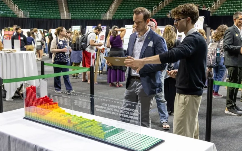 A graphic design student talks with another person about lego design that has a gradient of red to yellow to green in a glass casing, set up in a room with other participants and exhibits.