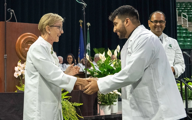 Two people in white coats shake hands during a graduation ceremony with flowers lining the stage.