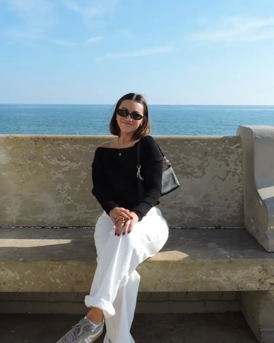 Woman wearing sunglasses, a black top and white pants sits on a stone bench overlooking a blue ocean under a clear sky.