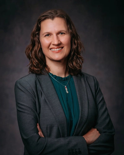 Studio portrait of a smiling woman with shoulder-length wavy brown hair, wearing a gray blazer over a dark teal blouse with a silver pendant necklace, standing with arms crossed against a dark textured background.