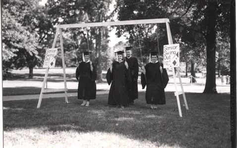 This photograph shows four senior students smiling by the swings in their commencement robes. The signs read "Senior Swing Out June 3 7:30pm".