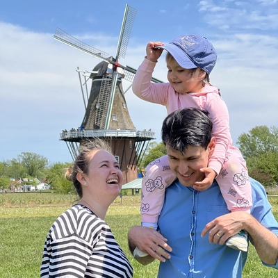 A photo outside of a family- a woman with brown hair in a white and black striped shirt, a man in a blue button down and their daughter on his shoulders in all pink standing in front of a windmill.