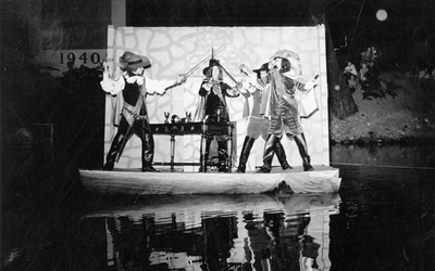 Four students in costume, holding up their swords towards the sky, while riding on a parade float in the Red Cedar River.
