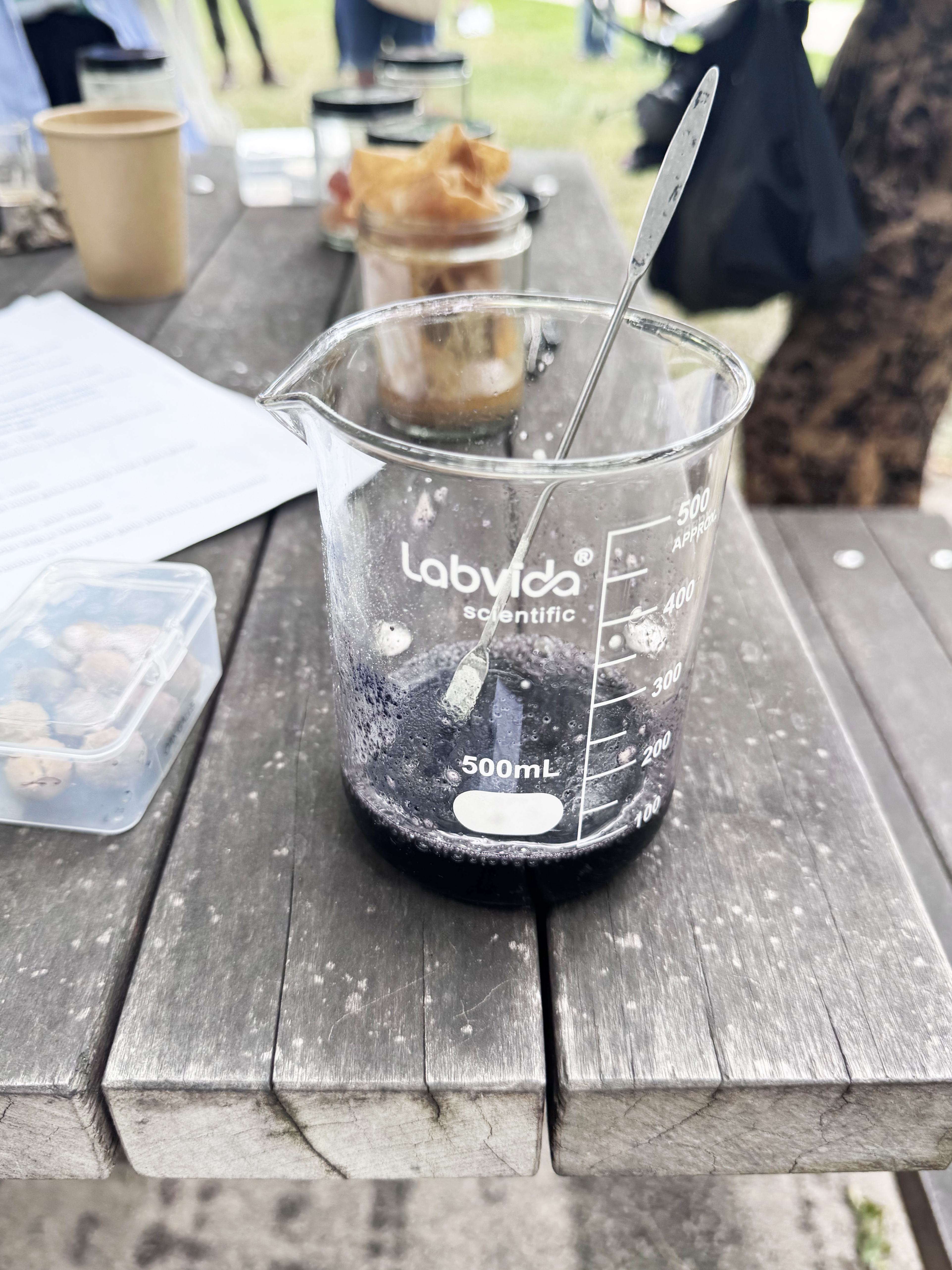 Clear 500-milliliter beaker containing dark liquid with a metal stirring tool rests on a wooden table alongside other containers and materials.