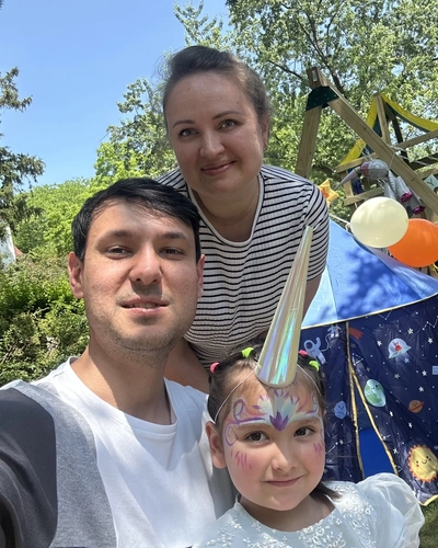 A photo of a woman in a black and white stripe shirt standing behind a man in a white and gray shirt with a young girl with colorful facepaint. they are sitting outside.